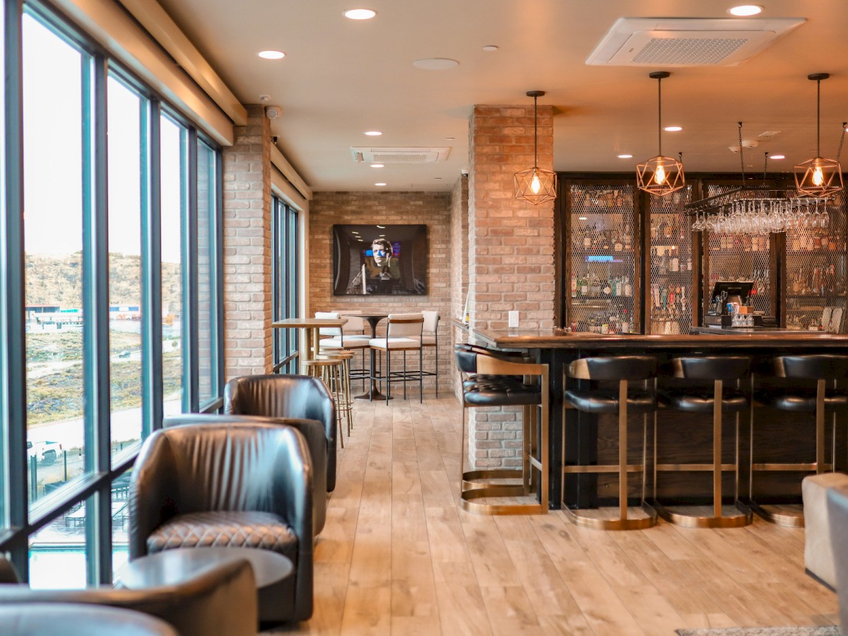 A modern lounge bar with leather chairs, high-top tables, large windows, and a well-stocked bar area. Cozy and inviting atmosphere.