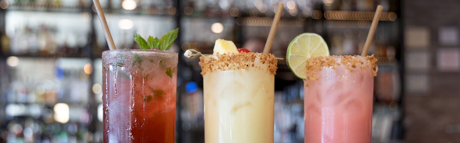 Three colorful cocktails in tall glasses, garnished with mint, lime, and cherries, on a bar counter with a blurred background.