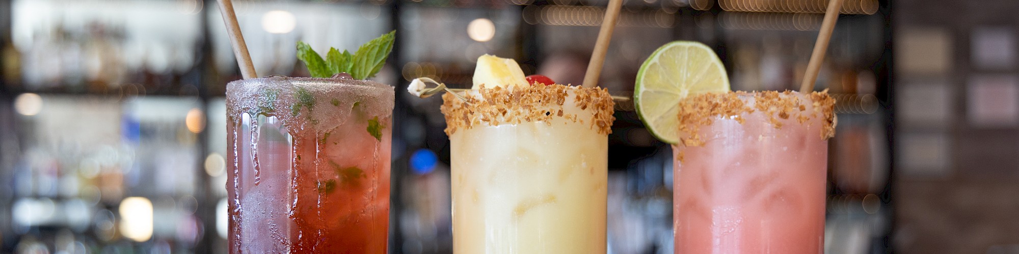 Three colorful cocktails in tall glasses, garnished with mint, lime, and cherries, on a bar counter with a blurred background.