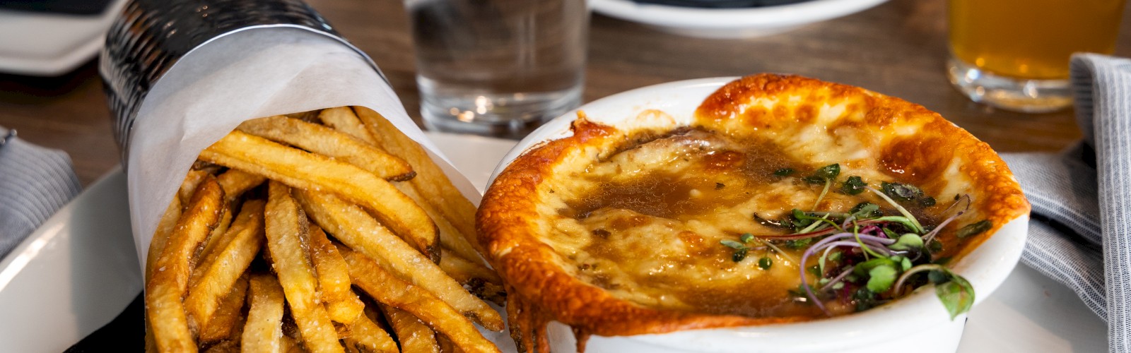 The image shows a meal with fries wrapped in paper and a bowl of baked soup topped with cheese, garnished with herbs on a white plate.