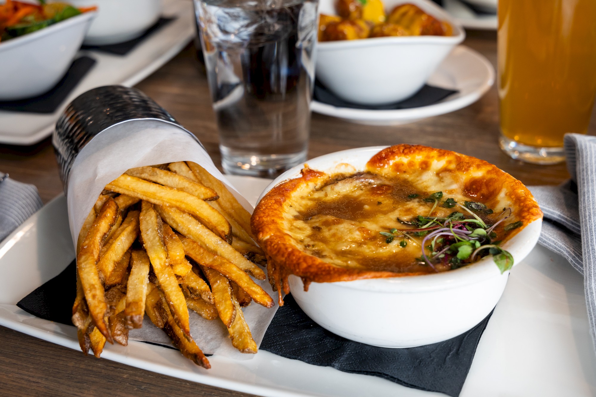 The image shows a meal with fries wrapped in paper and a bowl of baked soup topped with cheese, garnished with herbs on a white plate.