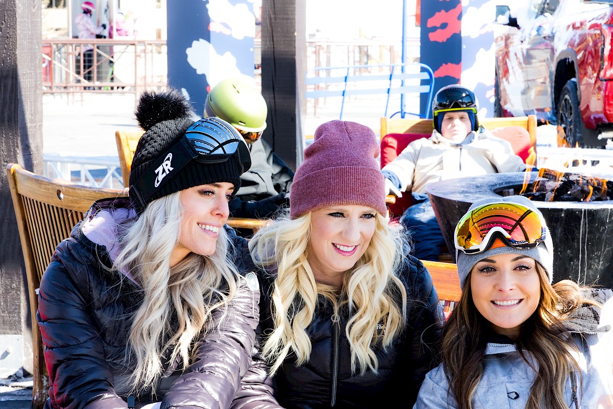 Three people in winter gear smiling outdoors, with ski goggles and beanies, enjoying a snowy setting. Others are in the background.