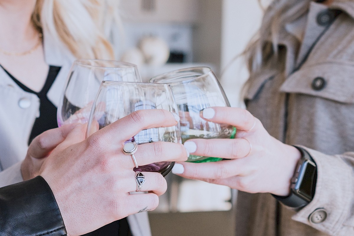 Three people are toasting with drinks, holding stemless glasses, and wearing casual clothing and accessories like rings and a smartwatch.
