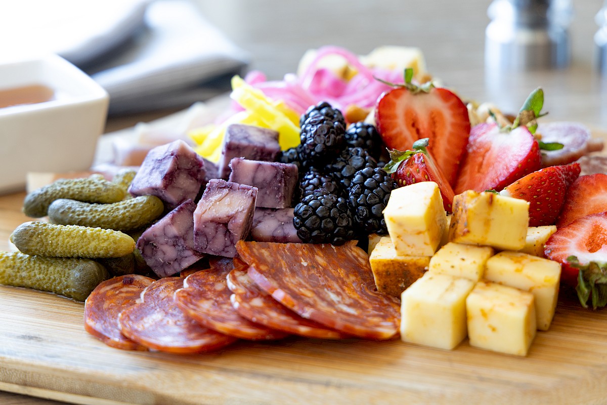 A charcuterie board with sliced salami, cheese cubes, berries, strawberries, pickles, and more, arranged on a wooden surface.