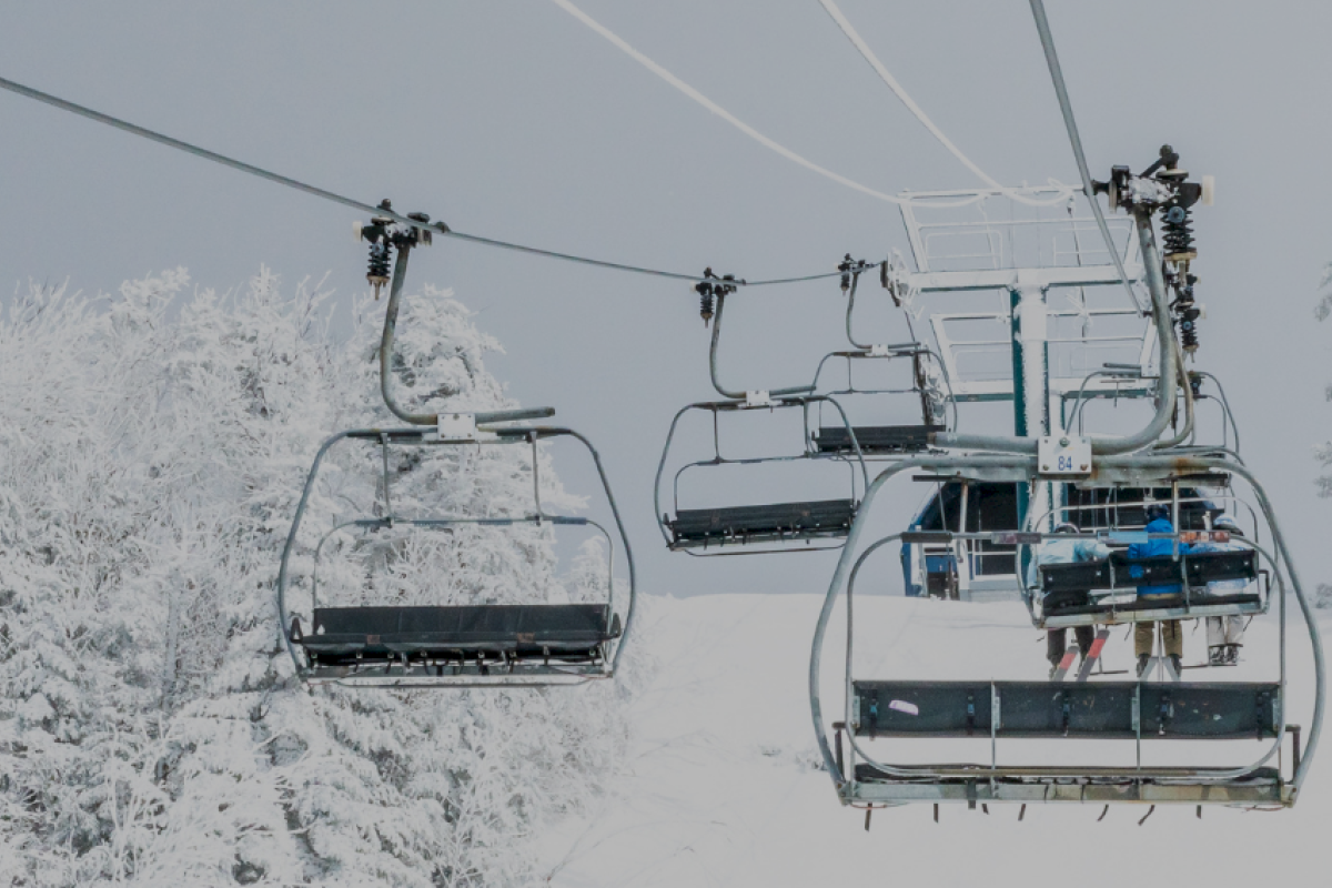 Empty ski lift chairs are suspended above a snowy landscape with snow-covered trees in the background.