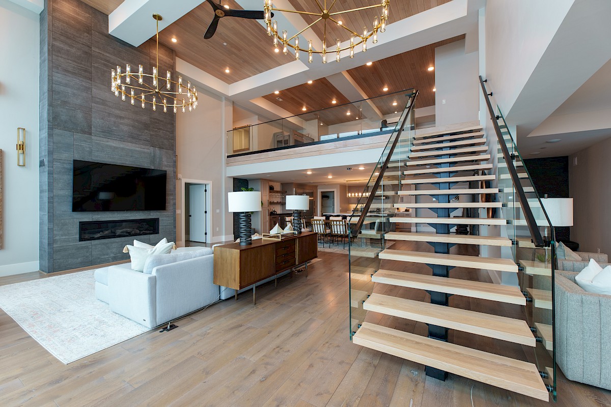 A modern, spacious living room with a high ceiling, chandeliers, a large staircase, and a mix of wooden and glass elements.