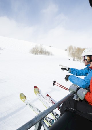 People sitting on a ski lift, wearing helmets and winter clothing, overlooking a snowy mountain with skiers below.