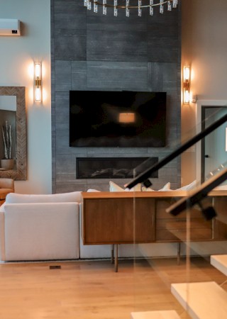 A modern living room with large windows, a TV, fireplace, and cozy seating, featuring a staircase and warm lighting on the walls.