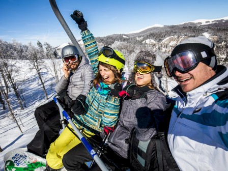 A group of four people in winter gear on a ski lift, excitedly posing for a selfie against a snowy mountain backdrop.