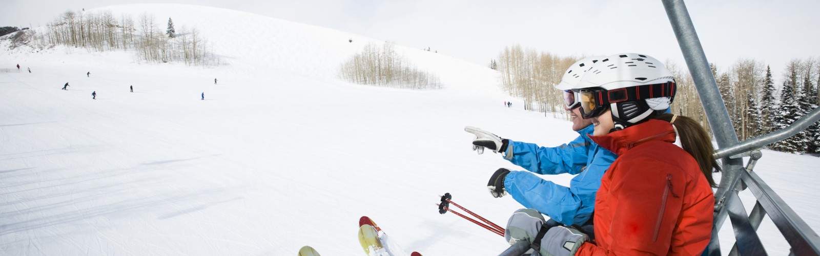 Skiers in helmets and gear riding a ski lift, pointing at the snowy slopes and distant skiers, with a clear sky above.