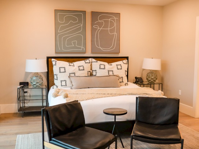 A modern bedroom with a double bed, abstract artwork, two chairs, side tables with lamps, and a small round table in front.