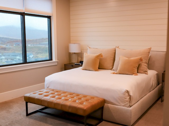 A cozy bedroom with a large bed, beige pillows, a tufted bench, lamp, and a window showing an outdoor view.