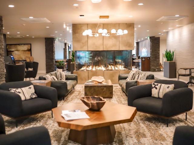 A modern lounge features plush chairs, a central wood table, and a fireplace, creating a cozy and inviting atmosphere.