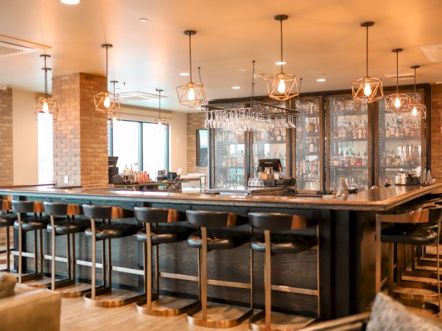 The image shows a modern bar with high stools, pendant lighting, and a well-stocked back shelf, creating a cozy, inviting atmosphere.
