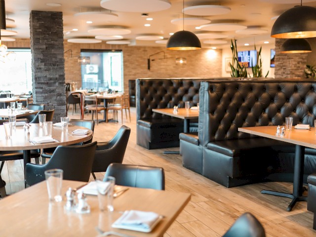 The image shows a modern restaurant interior with wooden tables, black leather booths, and ceiling lights, creating a cozy atmosphere.
