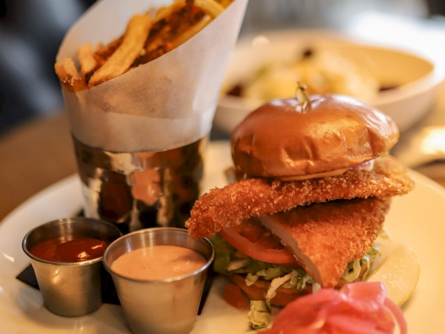 The image shows a burger with lettuce and tomato, fries in a metal cup, two dipping sauces, and pickles on a plate.