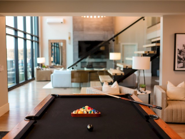 A modern living room with a pool table, couches, and a TV, featuring large windows and contemporary decor.
