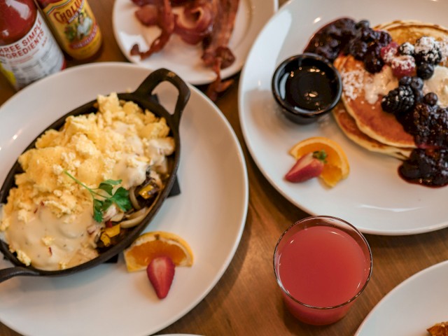 Plates of breakfast foods: pancakes with berries, an omelet with potatoes, a skillet dish, and a drink are on a wooden table.