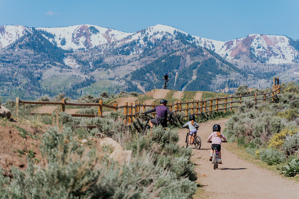 Four children and a man bike on a trail in a mountainous landscape with snow-capped peaks in the background.
