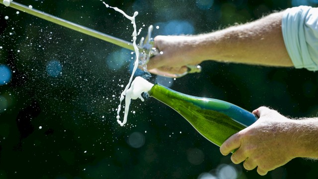 A person is using a sword to open a champagne bottle, with foam and liquid spraying, in a practice known as sabrage.