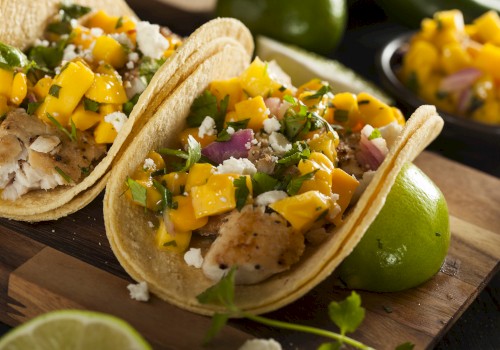 Two tacos with fish, mango salsa, cilantro, and cheese on a wooden board with limes. A dish with extra mango salsa is in the background.