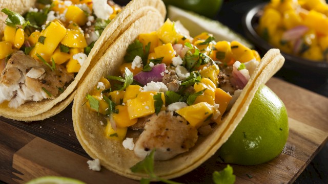 Two tacos with fish, mango salsa, cilantro, and cheese on a wooden board with limes. A dish with extra mango salsa is in the background.