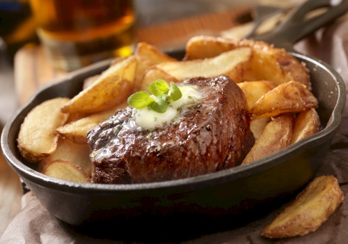 A sizzling steak topped with herb butter and served with potato wedges in a cast iron skillet, garnished with fresh herbs.