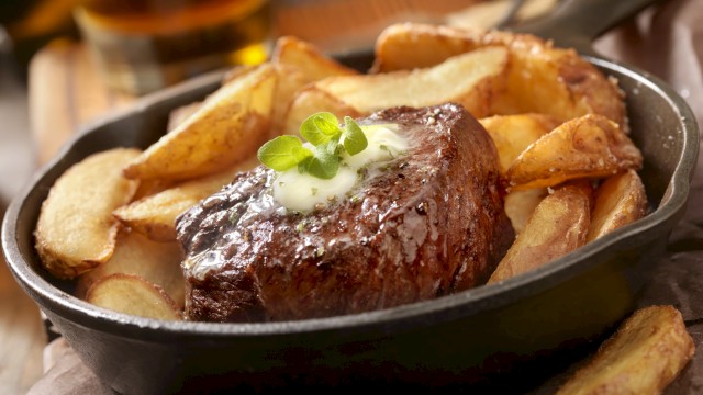 A sizzling steak topped with herb butter and served with potato wedges in a cast iron skillet, garnished with fresh herbs.