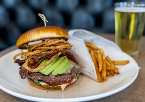 A hamburger with avocado, onion rings, fries on the side, and a glass of beer in the background on a wooden table.