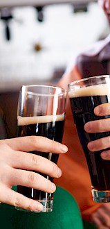 Deux personnes trinquent avec des verres de bière brune, l'une d'elles porte un trèfle, un drapeau irlandais drapé autour d'elles, suggérant une ambiance festive.