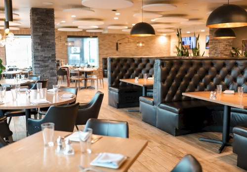 The image shows an empty modern restaurant with black chairs, wooden tables, leather booths, and warm lighting.