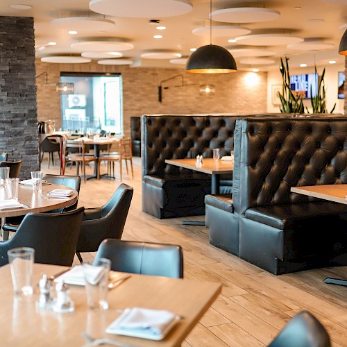 The image shows an empty modern restaurant with black chairs, wooden tables, leather booths, and warm lighting.