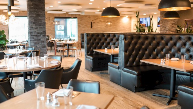 The image shows an empty modern restaurant with black chairs, wooden tables, leather booths, and warm lighting.