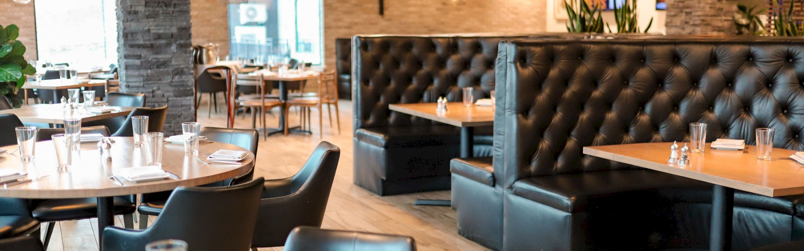 The image shows an empty, modern restaurant with wooden tables, black chairs, leather booths, pendant lighting, and a warm, inviting ambiance.