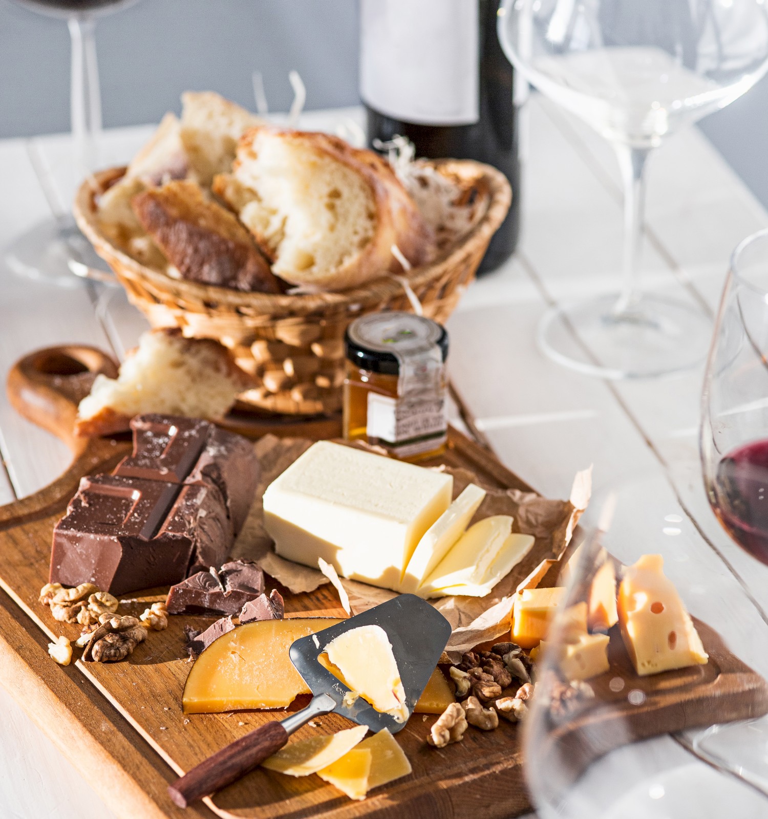 A cheese platter with assorted cheeses, nuts, and dark chocolate, alongside red wine, bread, and wine glasses on a wooden table.