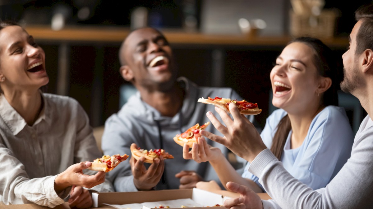 Four people are joyfully eating pizza together, sharing laughter around a table.