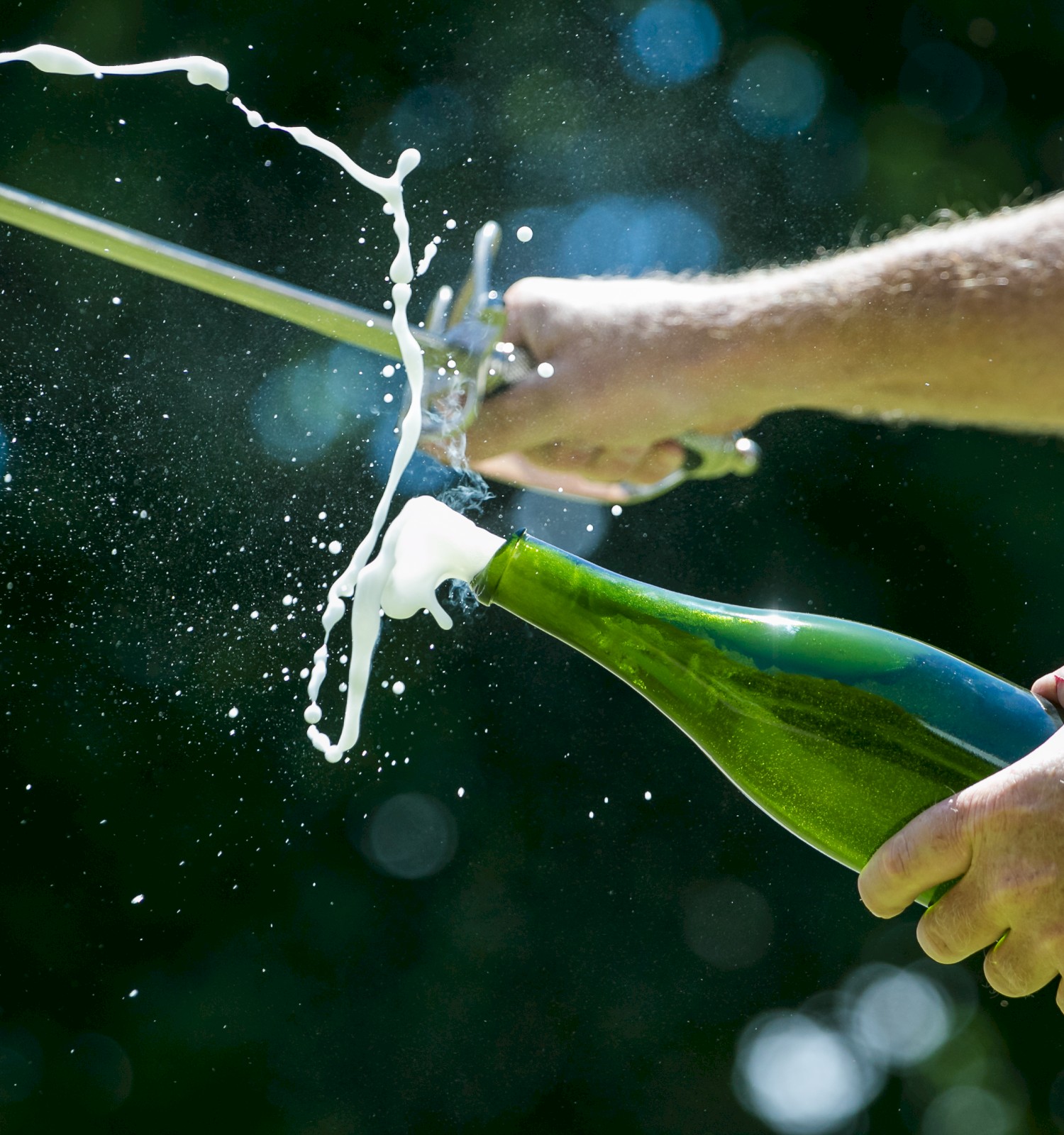 A person is using a sword to open a champagne bottle, creating a splash of foam and liquid in a green outdoor setting.