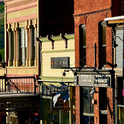 A colorful street with historic buildings, various store signs, and a decorative flag. Bright sunlight highlights the architectural details.