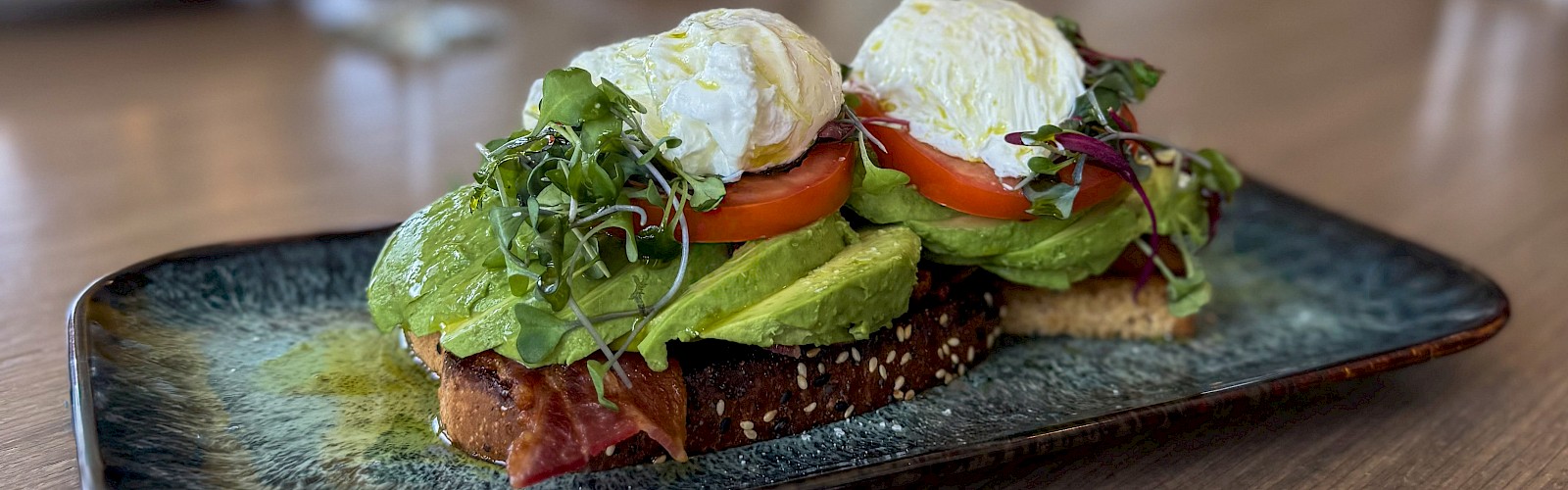 A plate of avocado toast topped with tomatoes, microgreens, poached eggs, and drizzled with oil, served on a dark ceramic dish.