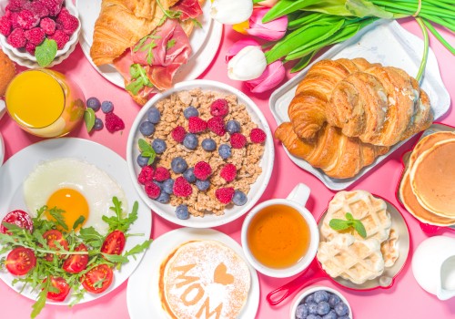 A colorful breakfast spread featuring pancakes, waffles, croissants, fruit, eggs, and flowers arranged on a pink surface.
