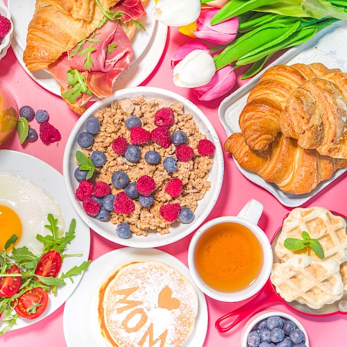 A colorful breakfast spread featuring pancakes, waffles, croissants, fruit, eggs, and flowers arranged on a pink surface.