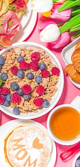Een kleurrijke brunch met croissants, pannenkoeken, granola, ei met groenten, bessen, sap en bloemen op een roze achtergrond.