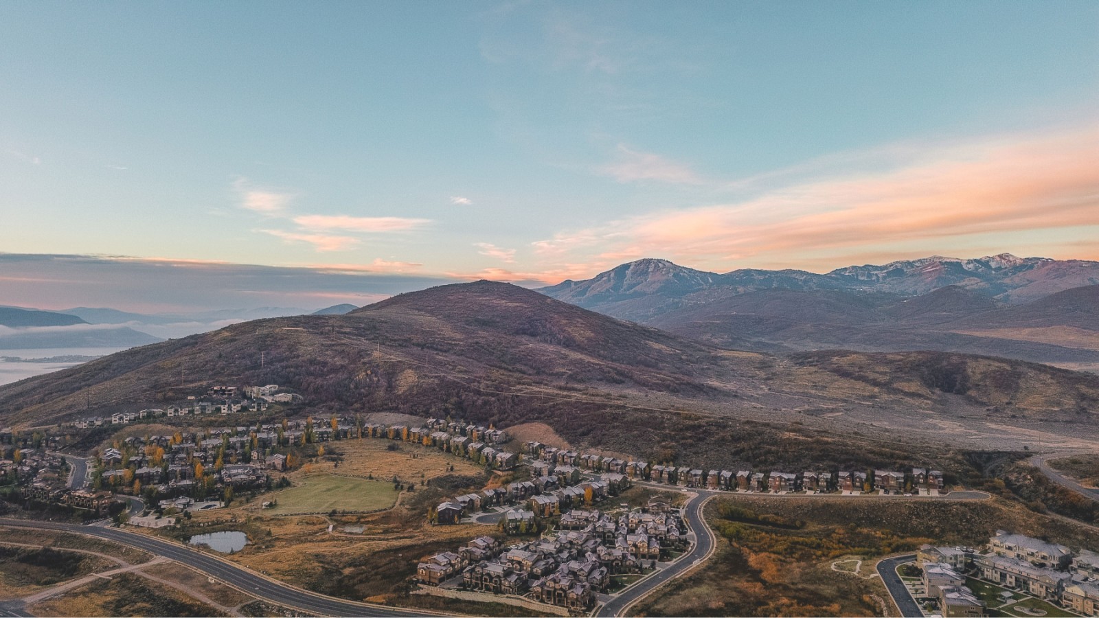 Aerial view of a residential area with houses nestled among hills and mountains under a partly cloudy sky.