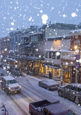 A snowy street scene with cars, buildings, and falling snow, creating a wintry atmosphere in an urban setting.