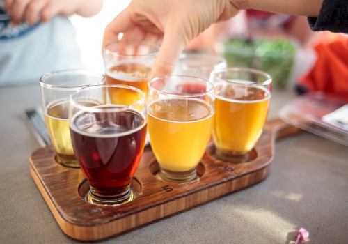 A hand reaches for one of several glasses of different colored beverages on a wooden tray, likely a beer flight, arranged on a table.