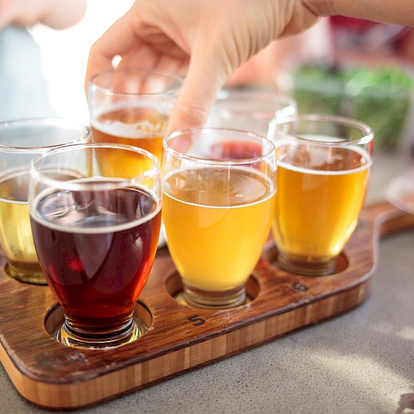 A hand reaches for one of several glasses of different colored beverages on a wooden tray, likely a beer flight, arranged on a table.