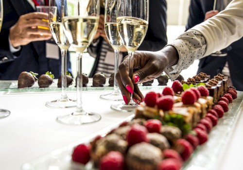 A hand reaches for desserts on a table with elegant glasses of champagne and people mingling in the background, showcasing a social gathering.
