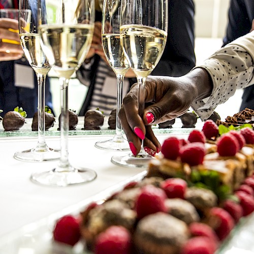 A hand reaches for desserts on a table with elegant glasses of champagne and people mingling in the background, showcasing a social gathering.