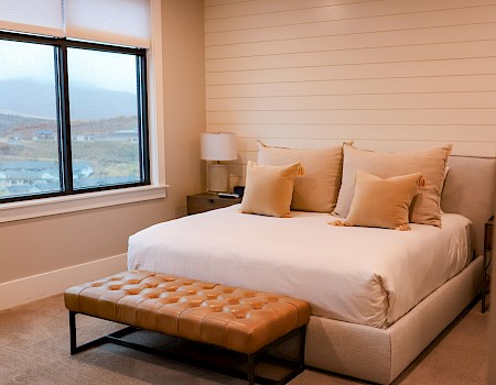 A cozy bedroom with a large bed, beige pillows, a tufted bench, nightstand with a lamp, and a window with a scenic view.
