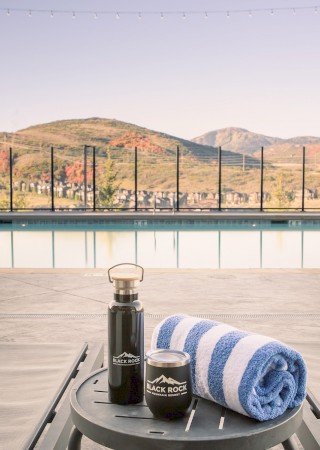 A poolside setup with a towel, water bottle, and cup on a table, against a scenic mountain background under string lights.